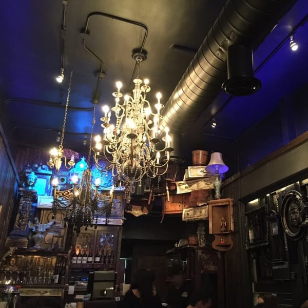 Ornate chandeliers illuminate a dark, eclectic interior with decorative objects and blue lighting.