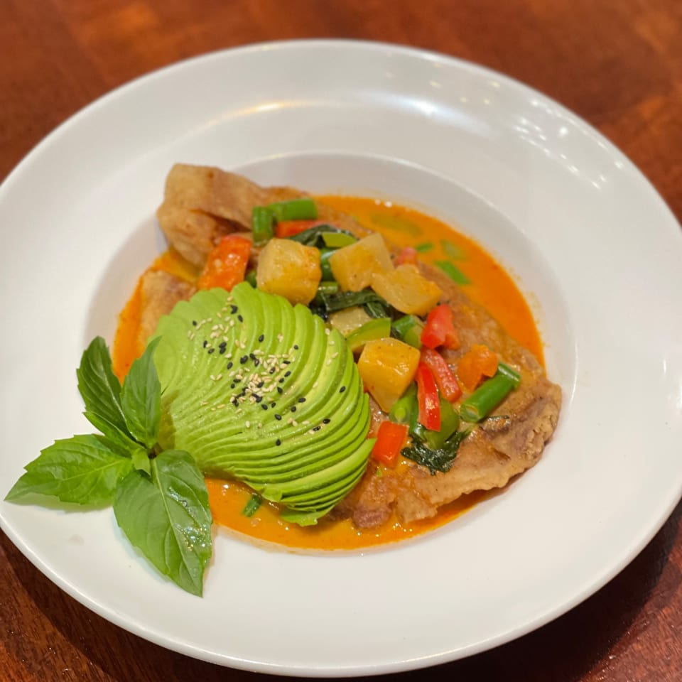 Thai red curry with fish, vegetables, and sliced avocado, garnished with basil and sesame seeds.