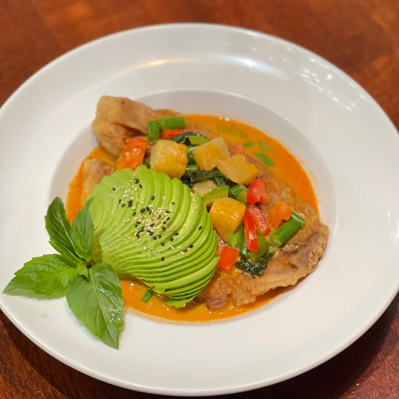 Thai red curry with fish, vegetables, and sliced avocado, garnished with basil and sesame seeds.