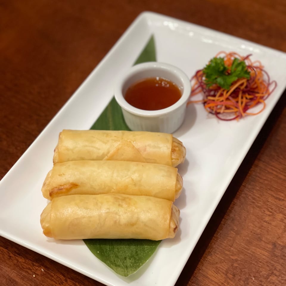 Three golden-brown Thai spring rolls on a white plate with dipping sauce and shredded vegetables.