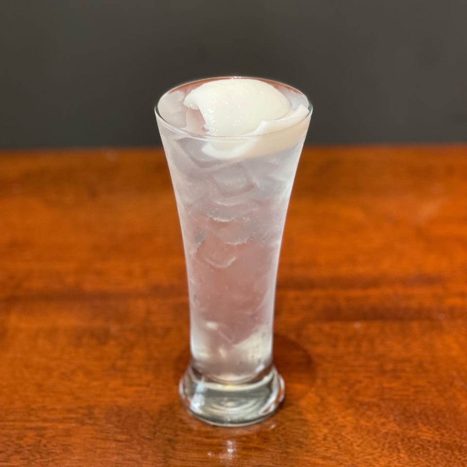 A tall glass of iced lychee soda with whole lychee fruit on top, on a wooden table.