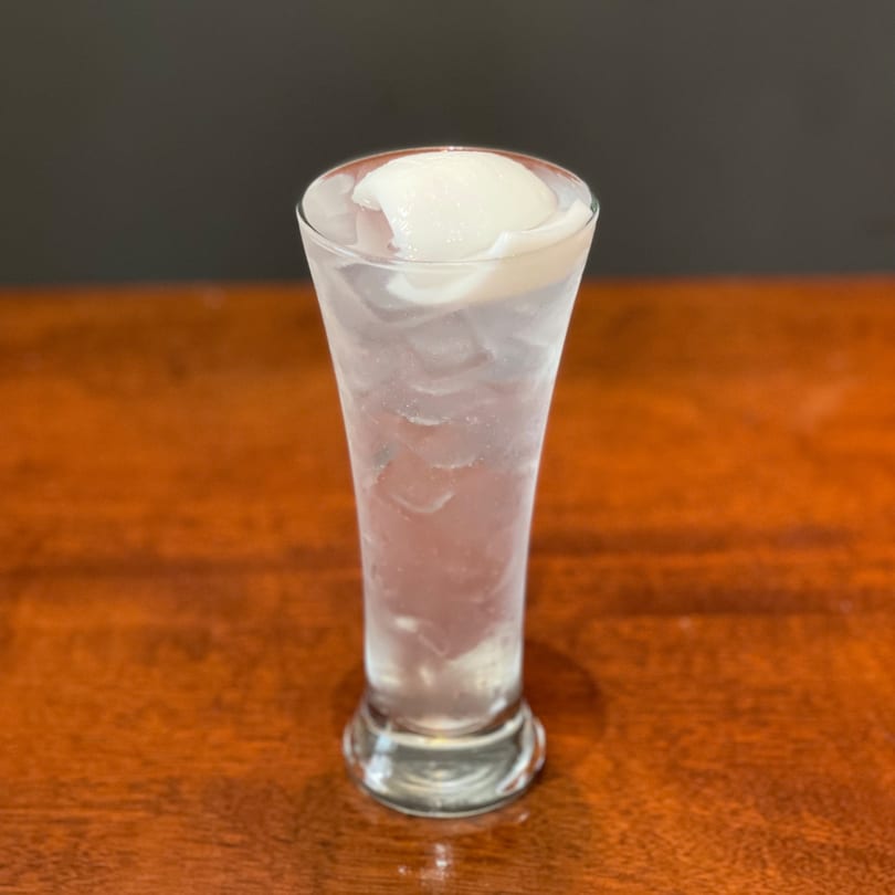 A tall glass of iced lychee soda with whole lychee fruit on top, on a wooden table.