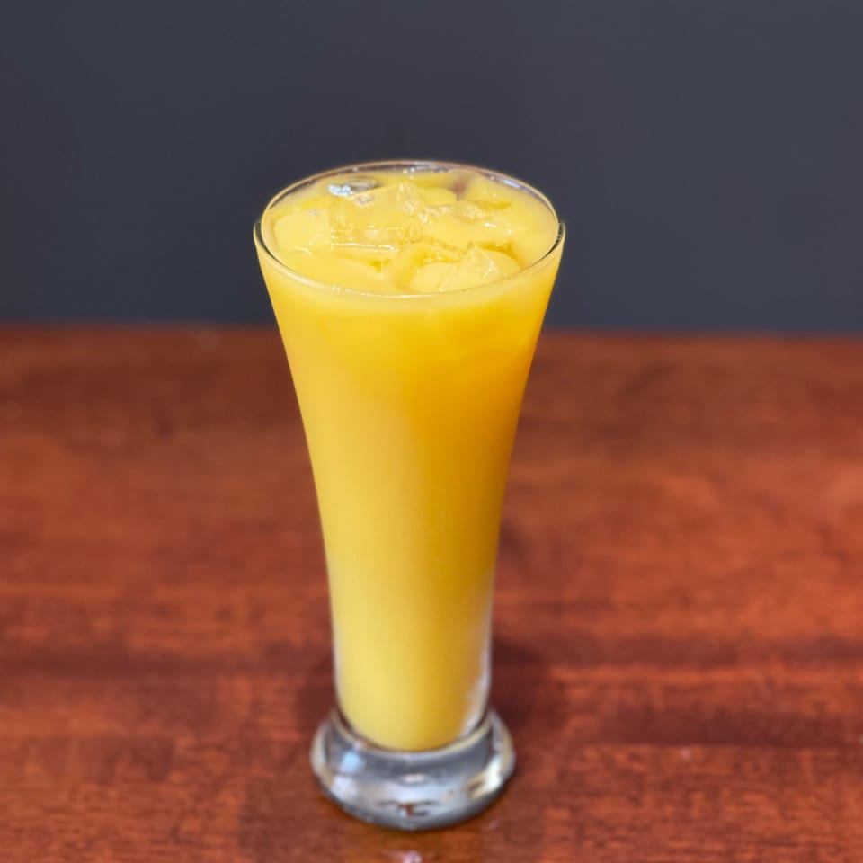 A tall glass of vibrant yellow mango juice with ice sits on a wooden table.