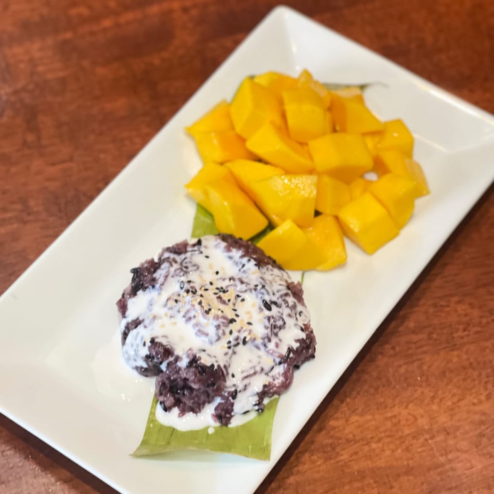 Black sticky rice with coconut milk and sesame seeds, served with fresh mango chunks on a white plate.