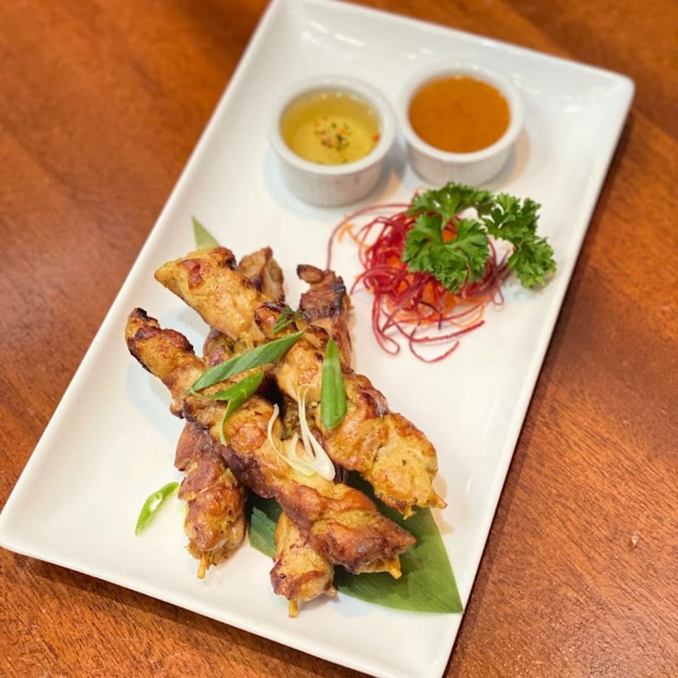 Thai chicken satay skewers with two dipping sauces, garnished with herbs and shredded vegetables.
