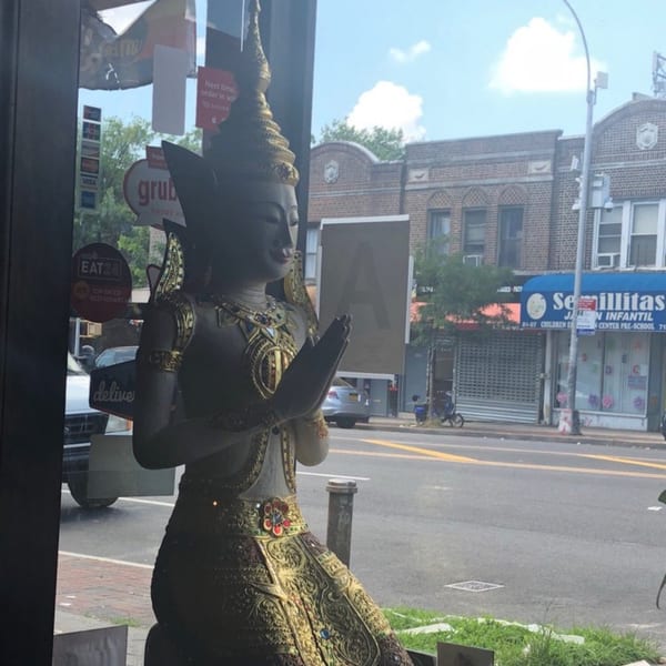 A golden Thai statue with hands pressed together in prayer, seen through a window overlooking a street.