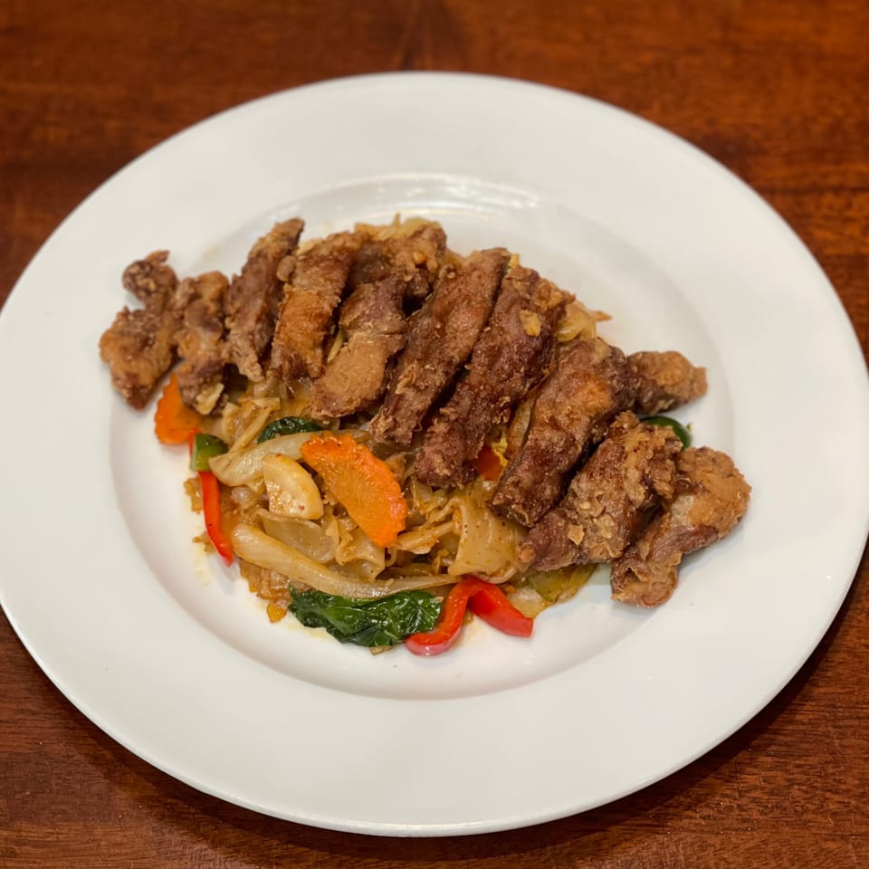 My Black Thai's crispy fried pork over stir-fried noodles with vegetables on a white plate.